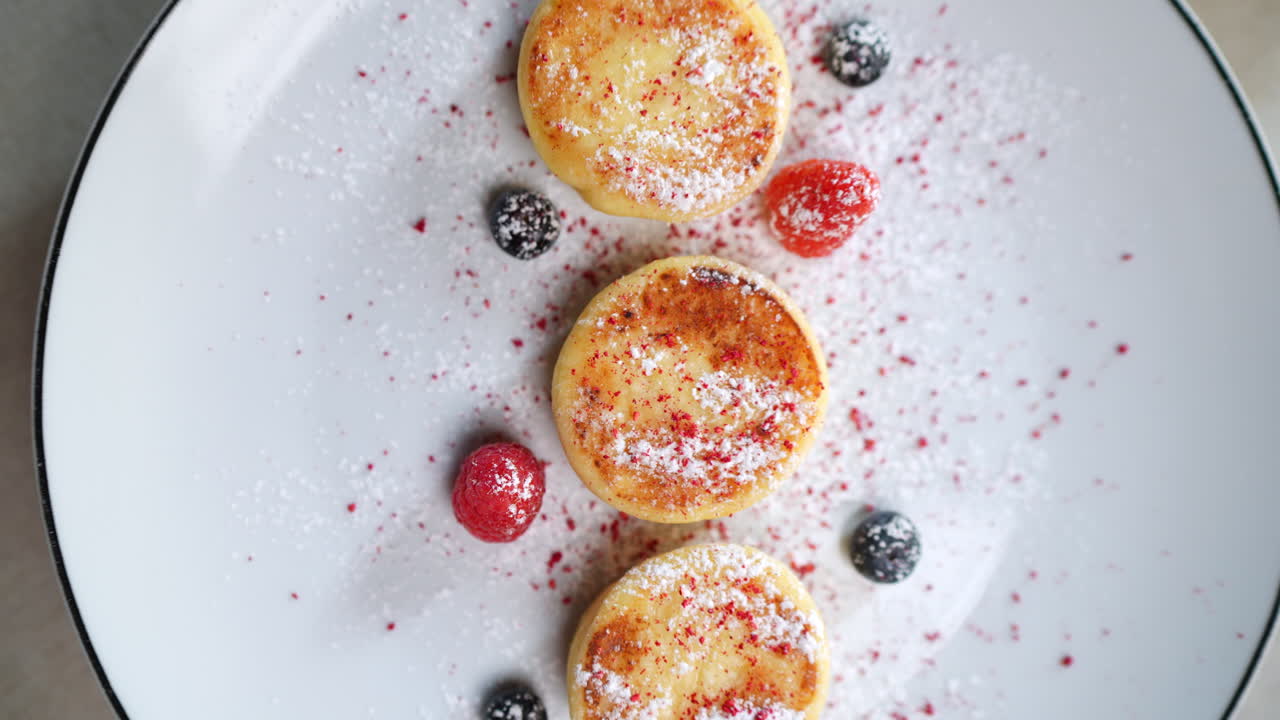 Small cottage cheese pancakes called syrniki, typical of Eastern European cuisine, served on a white plate, garnished with red fruits and blueberries, with powdered sugar and a fork on the side