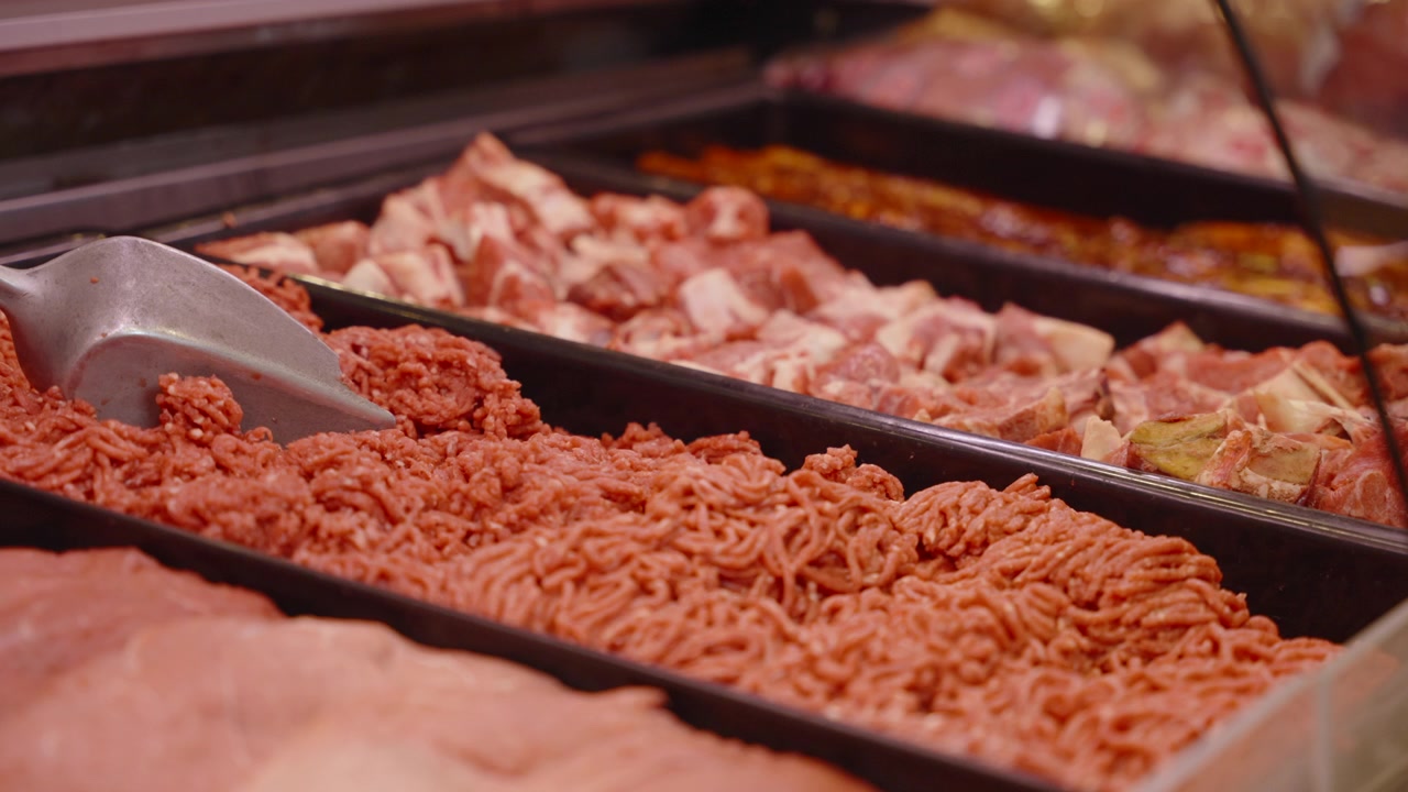 Carne macinata e tagli di macelleria assortiti disposti in vassoi di metallo sotto vetro, con un mestolo appoggiato nel banco frigo espositivo