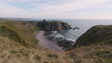 El-Castillo-De-Dunnottar-Se-Yergue-Orgullosamente-Sobre-Una-Roca-En-El-Fondo,-Mientras-Se-Desarrolla-Una-Hermosa-Playa-Con-Olas-Rompientes