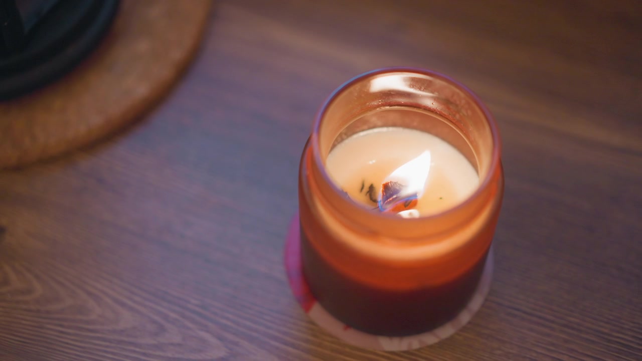 Nahaufnahme einer brennenden Kerze in bernsteinfarbenem Glas auf Holzoberfläche. Wachs schmilzt leicht, während die Flamme flackert und warmes Licht sowie zarte Schatten auf den Holzmaserungshintergrund wirft