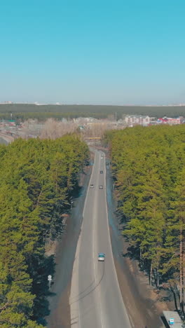 cars drive along road among evergreen forest against city silhouette on horizon under clear blue sky upper view