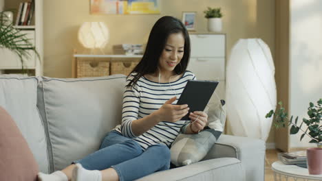 young woman watching something on the tablet and tapping on the screen sitting on a sofa in the living room