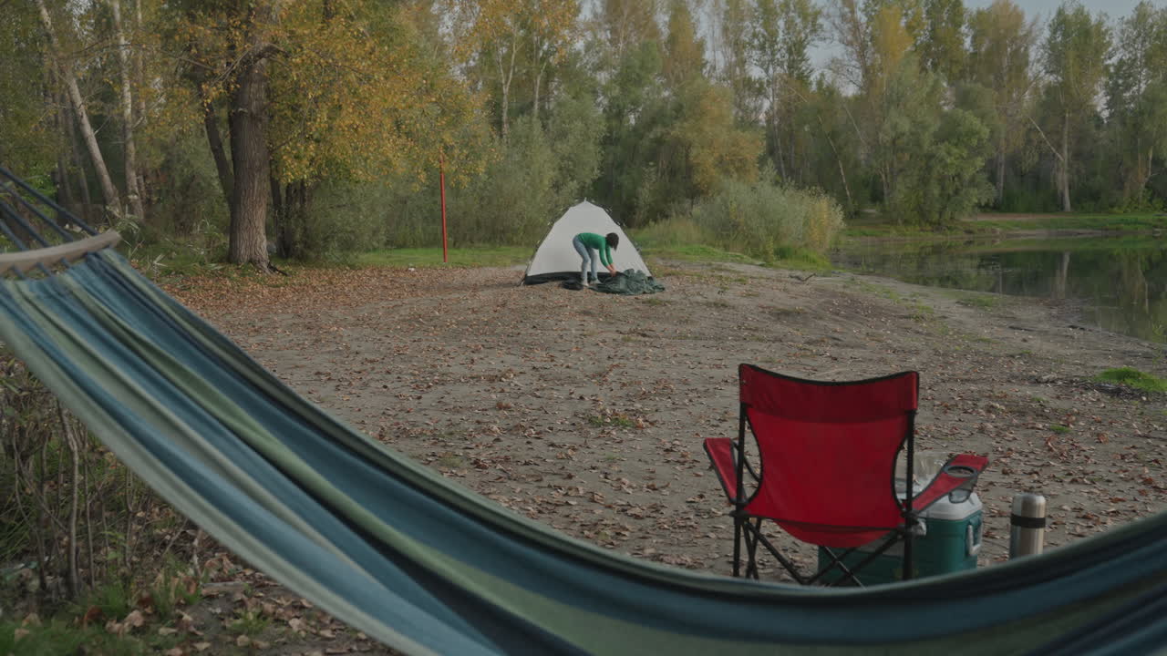 Opsætning af hængekøje og stol til camping ved søen. Teltet er slået op på den stenede strand, en termokande og et krus står ved siden af en foldestol. Roligt vand og gyldne træer i en stille efterårseftermiddag, ideel til fredfyldt ensomhed.