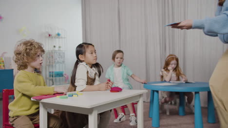 female teacher giving play dough to her pupils sitting at desk in montessori school