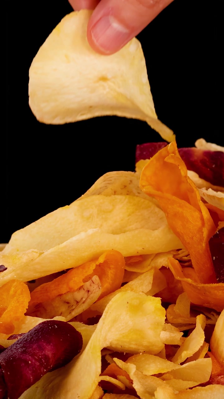 Hand selects a crispy root vegetable chip from assorted snack pile, brightly lit, close-up view