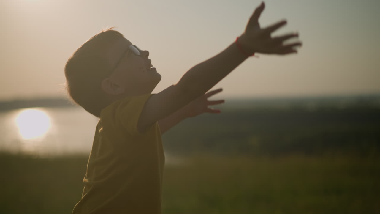 A little boy wearing a yellow shirt and glasses, joyfully reaches out to touch colorful bubbles floating gracefully in the air, with a serene sunset and a distant river in the background