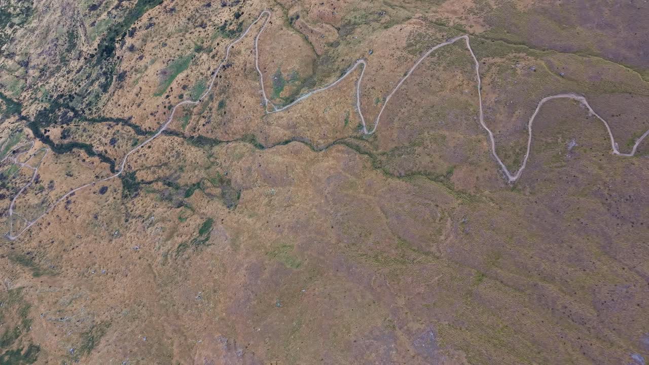 Drone in full top-down view advancing above the winding hiking trail of Roys Peak in Wānaka, revealing its curves and alpine landscape