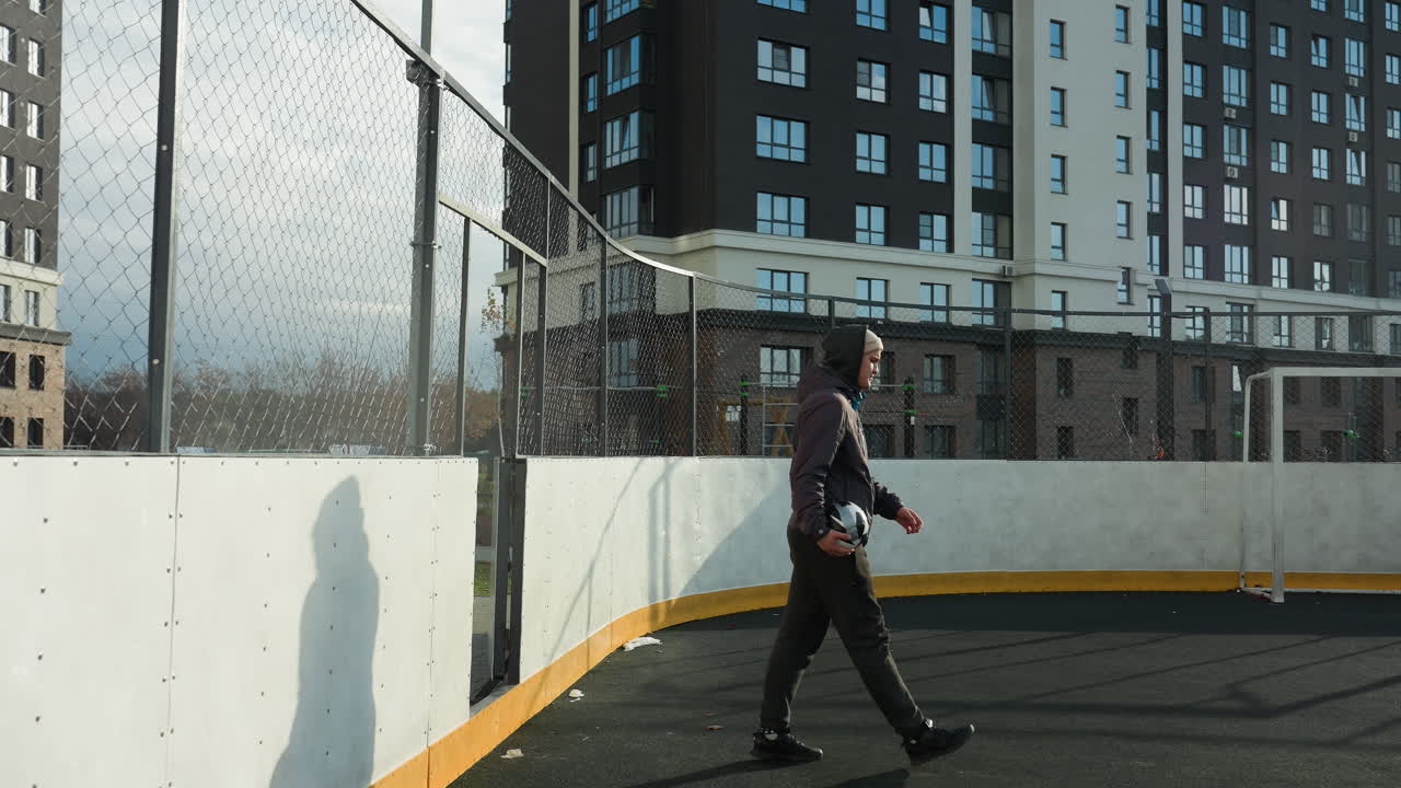 hombre con capucha y gorra de gorra entra en el campo de fútbol sosteniendo la pelota en el brazo derecho con la red de bares y edificios de oficinas en el fondo, mostrando el entorno deportivo urbano
