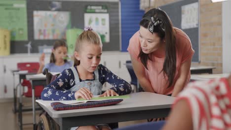 una feliz maestra diversa hablando con una niña en silla de ruedas en una clase de escuela primaria, cámara lenta