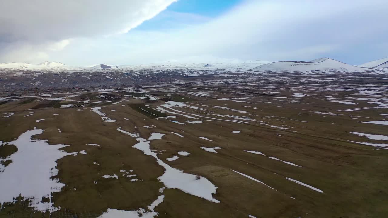 Høyfjellslette med spredt snø og fjerne fjell, drone stiger over vidåpent landskap, dramatisk himmel, natur i kaldt klima.