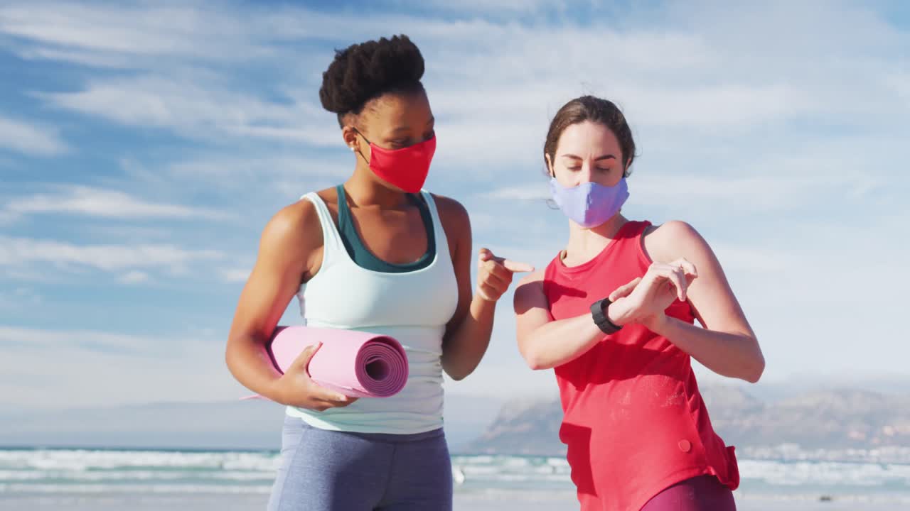 dos amigas diversas con máscaras faciales sosteniendo esteras de yoga en la playa