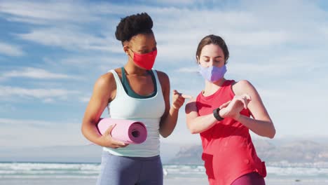 two diverse female friends wearing face masks holding yoga mats at the beach