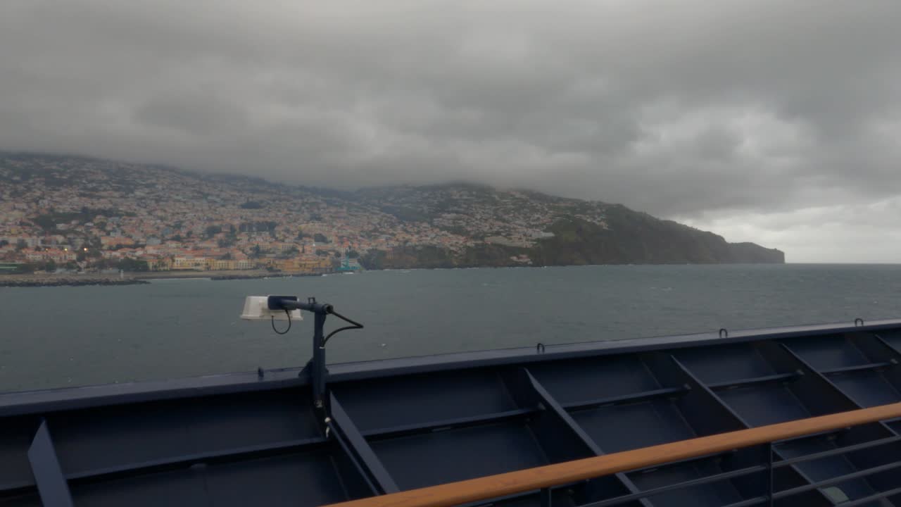 Vista dal ponte di una nave da crociera in avvicinamento a Funchal, Madeira, Portogallo. Cielo drammaticamente coperto sopra la città collinare e la costa rocciosa. Scena di viaggio marittimo suggestiva.