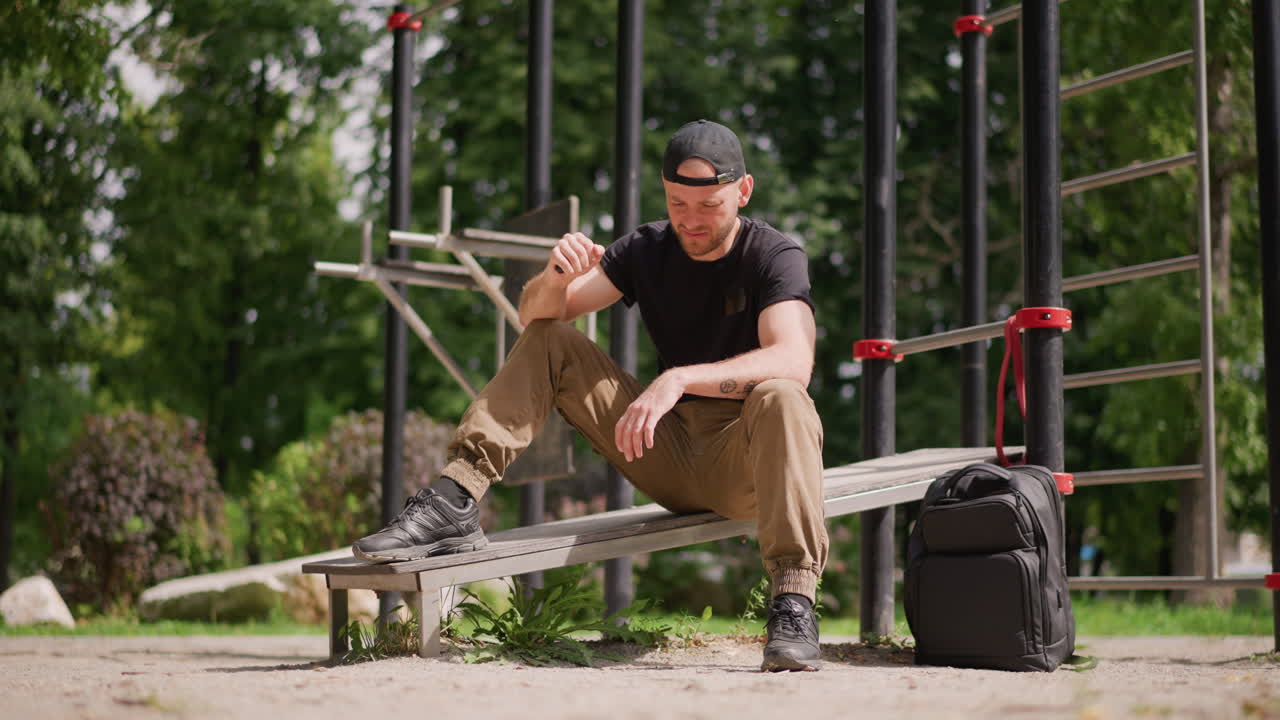 Park Rest Break By White Man, Seated Recovery On Metal Bench, Sipping Water And Catching Breath, Backpack At Side, Leafy Shade, Reflective Pause After Intense Calisthenics Session, Quiet Urban Green