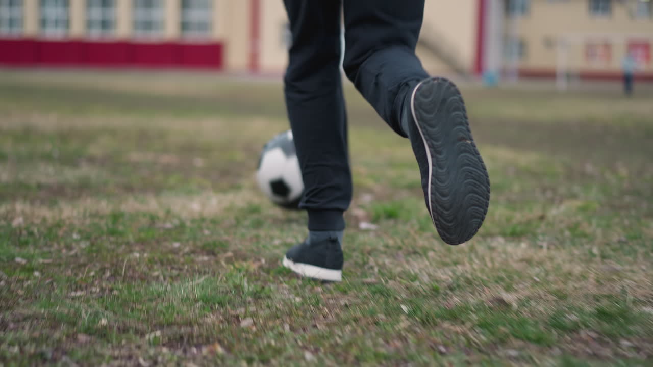 Close-up of someone in black pants and sneakers running with a soccer ball towards a goal post, with a blurred view of a person standing at the goal and buildings in the background