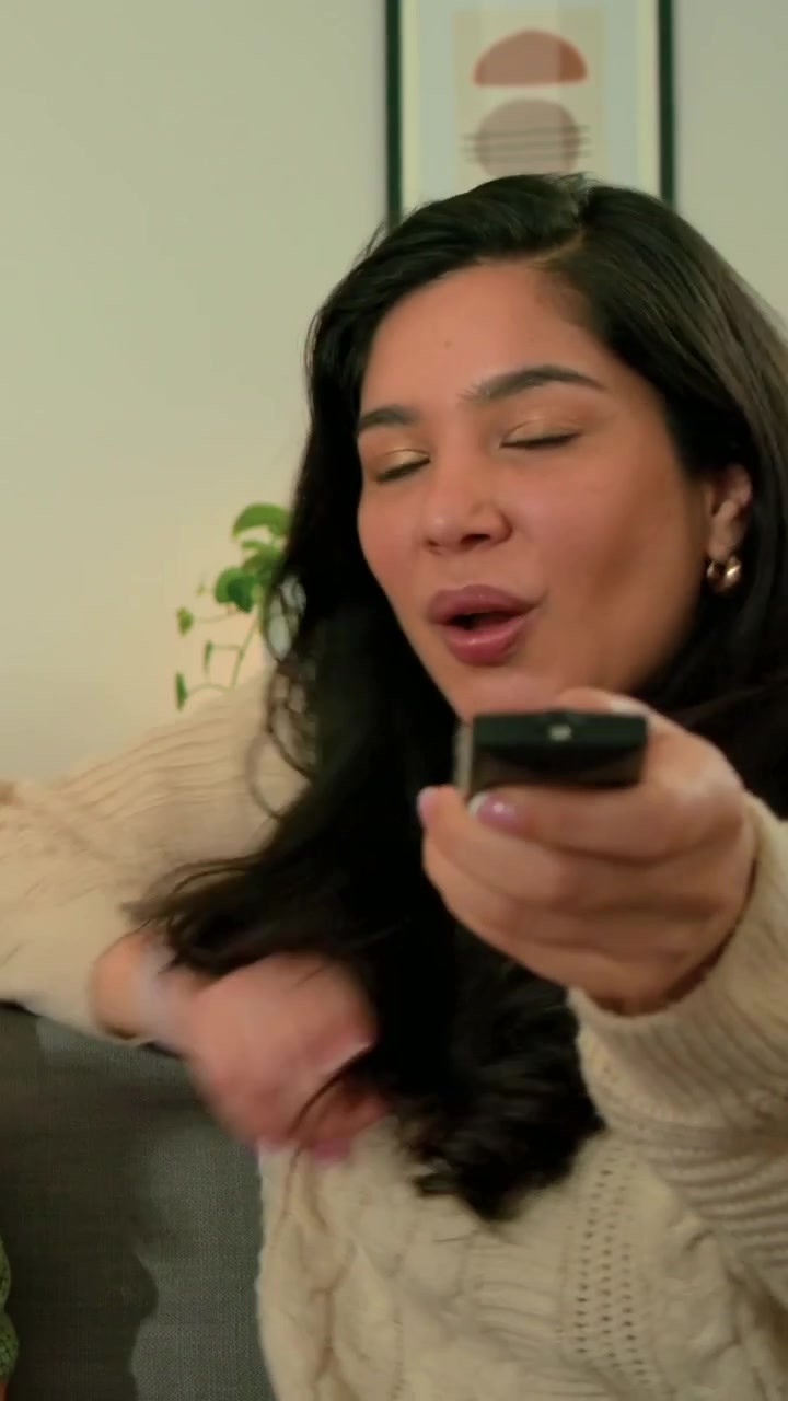 Vertikal video: Smilende kvinde tilbyder fjernbetjening, efter at en ven har talt på sofaen derhjemme, iført cremefarvet sweater.