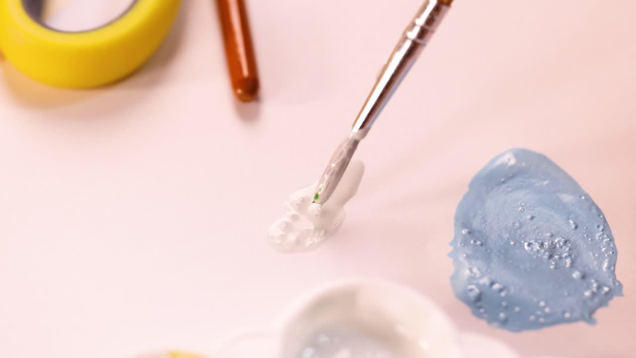 Hand mixes green and blue acrylic paint with brush on pastel workspace under soft lighting