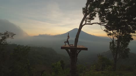 woman doing warrior pose on wooden platform with misty view of mount agung, dusk