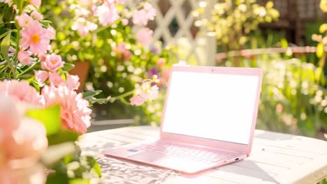 pink laptop on an outdoor table surrounded by blooming flowers in a sunny garden