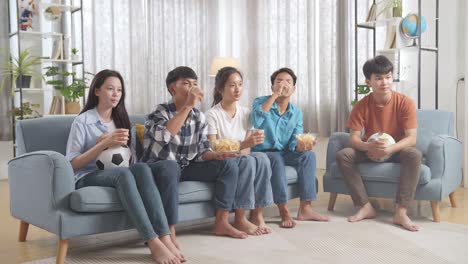 asian teenagers cheering, eating and drink while watching football game on tv at home