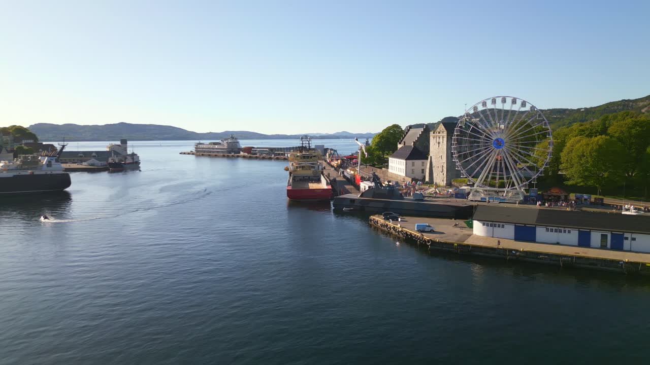 Aerial view of Bergen harbor, Norway, on a clear summer day, featuring docked boats and a lively carnival park with a Ferris wheel.