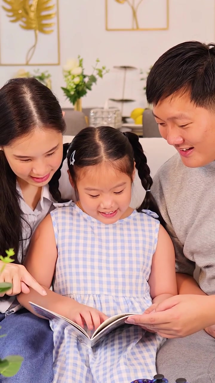 Cha mẹ và con gái mỉm cười đọc sách trong một phòng khách sáng sủa, ấm cúng với đồ ăn nhẹ
