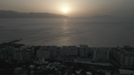 Drohnenaufnahme-über-Der-Stadt-Vlore,-Albanien,-Mit-Blick-Auf-Die-Gebäude-Und-Den-Hafen-Mit-Der-Fähre-Mit-Dem-Meer-Und-Dem-Strand-Darunter-Während-Des-Sonnenuntergangs-Am-Abend-Mit-Einem-Orange-Leuchtenden-Baumstamm