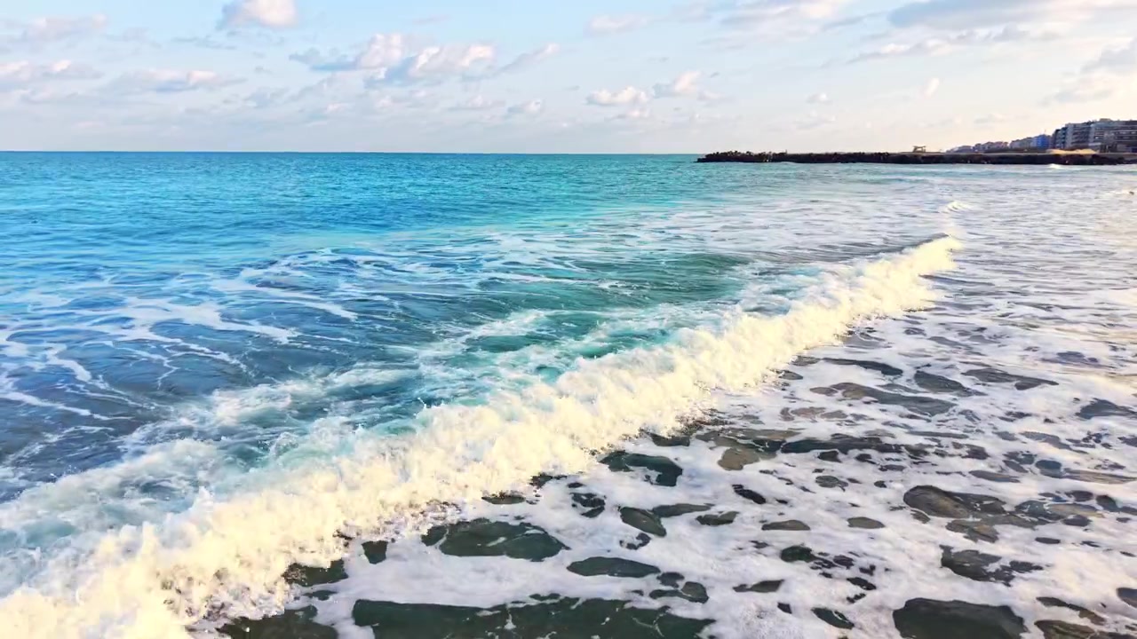 Schaumige Wellen rollen an den Strand. Türkises Meer, weißer Schaum und entfernte Küstenstadtsilhouette.