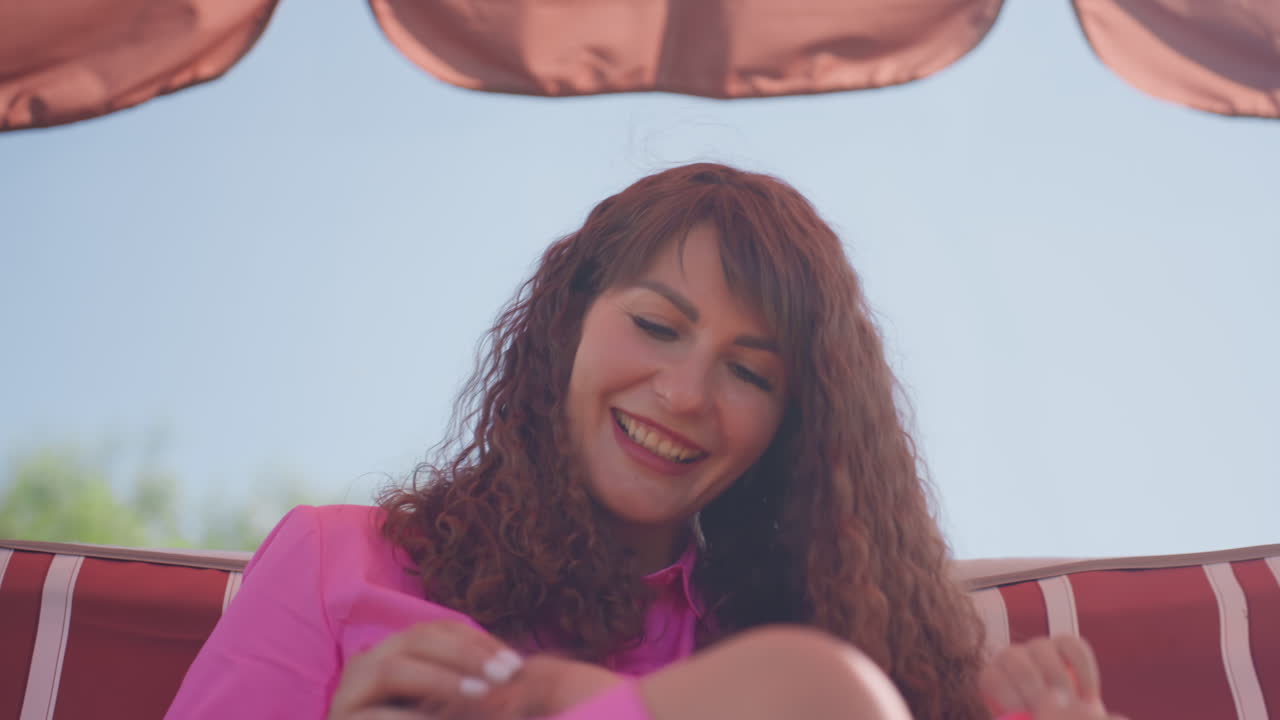 Woman Resting On Colorful Seat, Relaxed Woman In Pink Dress Sitting Peacefully On Outdoor Swing, Serene Lady In Vibrant Attire Peacefully Lounging On Striped Swing Beneath Lush Canopy In Garden