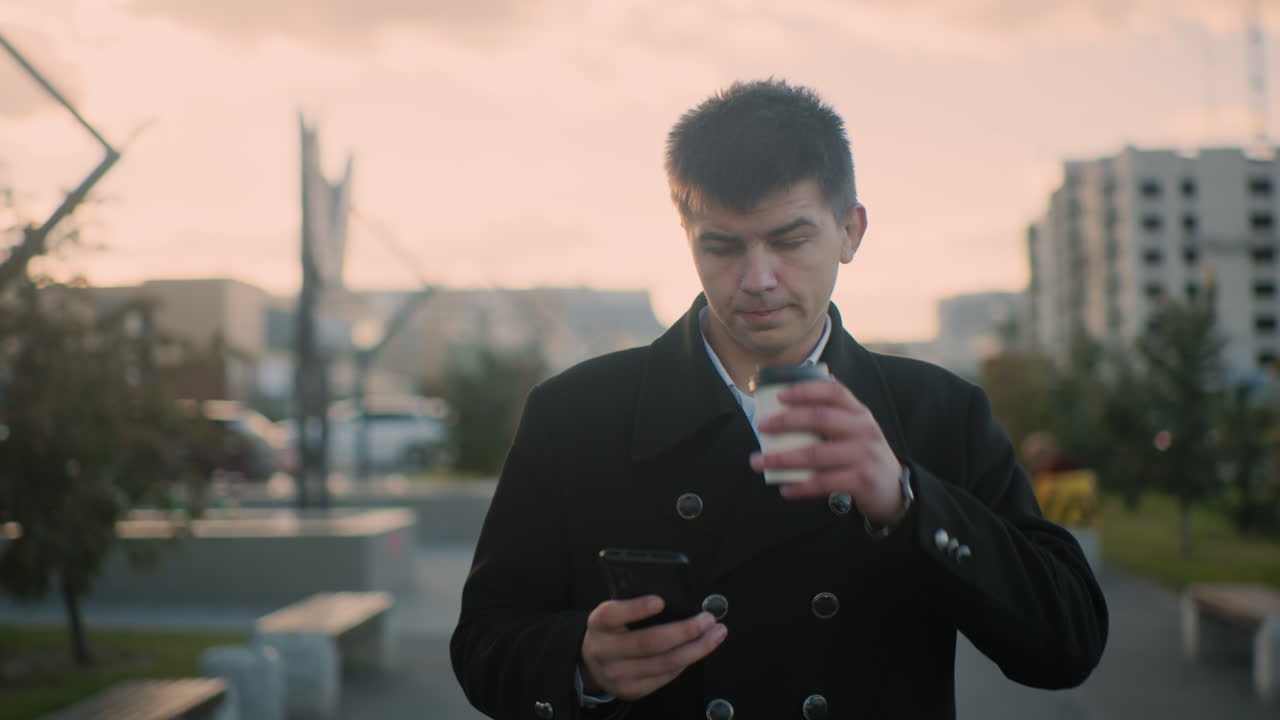 야외에서 커피를 마시고 도시 환경에서 휴대전화에 집중하고 녹색, 주차된 자동차, 움직이는 자동차, 그리고 배경에서 흐릿하게 는 사람들을 특징으로합니다.