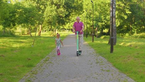 Senior-man-grandfather-tourist-riding-electric-scooter-with-granddaughter-child-girl-in-summer-park