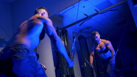 shirtless bodybuilder during workout in blue light. reflection in a mirror of a young bodybuilder training his biceps. muscular man exercising in the gym.