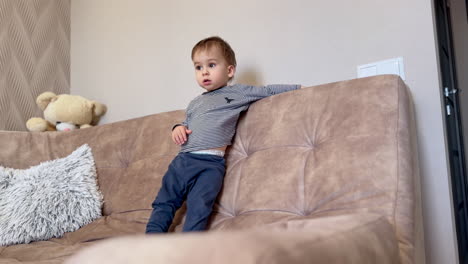 sweet little toddler standing on the sofa. baby waves his hands and smiles adorably. low angle view.