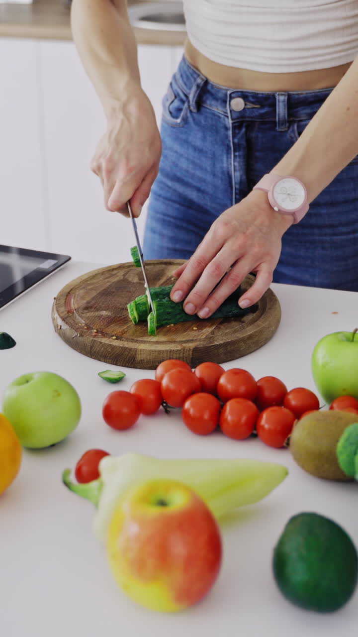 Frutta e verdura deliziose su un tavolo e una donna che cucina. La casalinga sta tagliando cetrioli verdi su un tagliere di legno per preparare un'insalata fresca in cucina. Video verticale.