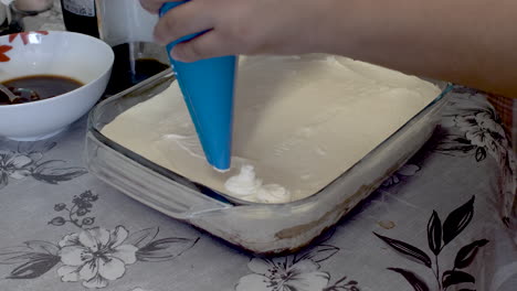 baker applying fresh cream using piping bag to tiramisu desert cake on kitchen table