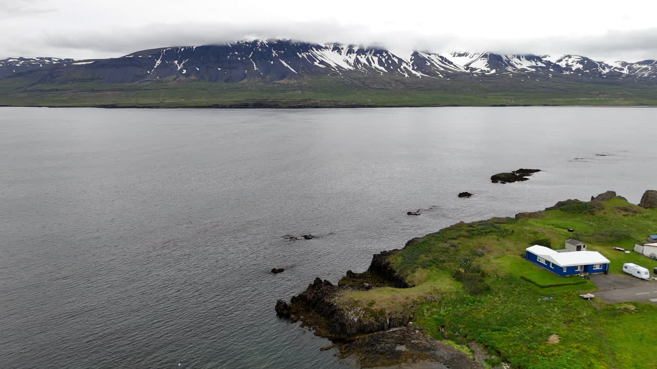 Aerial drone view of Vopnafjörður in Austurland, showing a dramatic coastline, dark cloudy skies, wide open fields, and the calm shoreline of this remote Icelandic region