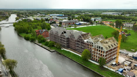 An-aerial-shot-of-a-construction-site-by-a-river-in-Elbląg,-with-partially-completed-buildings-and-a-construction-crane,-set-against-a-backdrop-of-green-areas-and-the-city-beyond