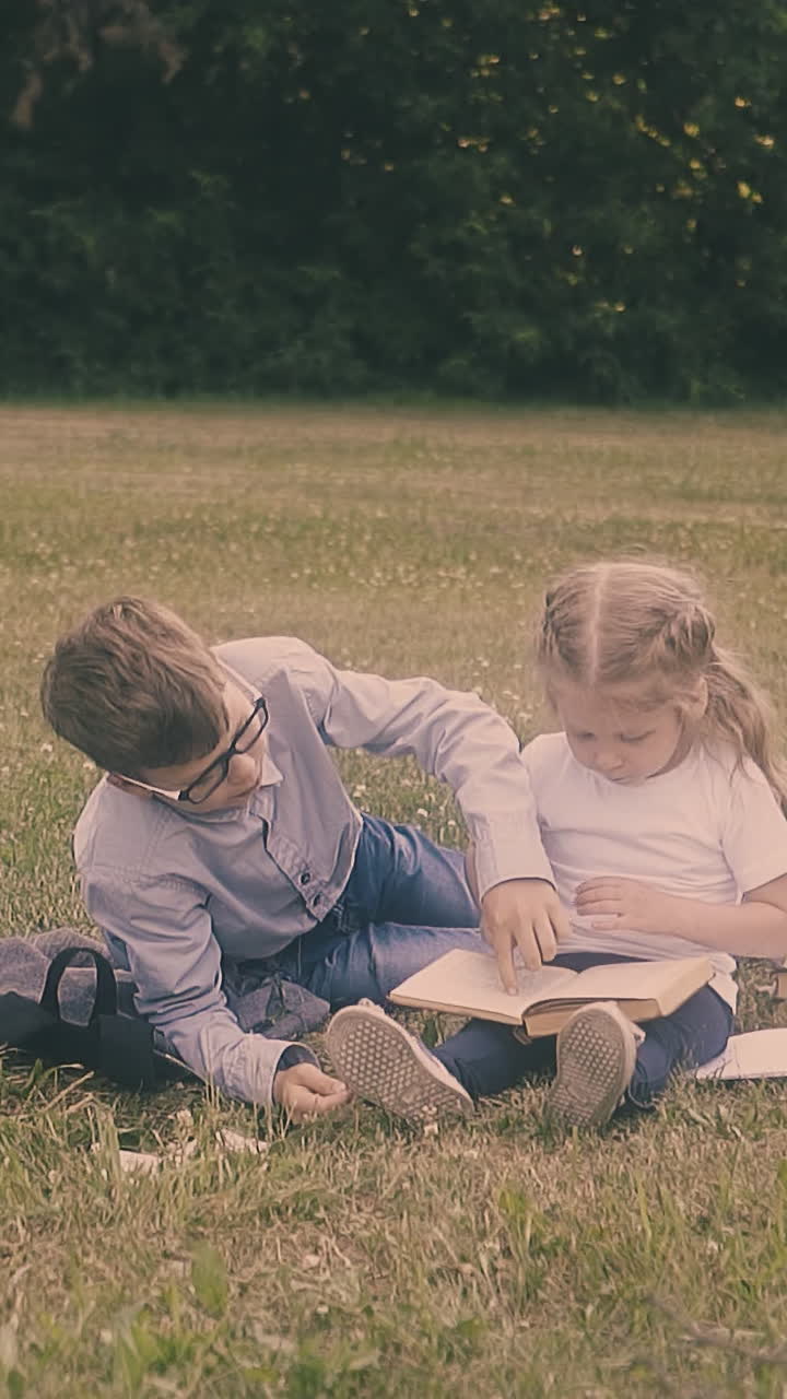 schoolboy in glasses explains math to small girl sitting near eating classmate on green lawn close view slow motion
