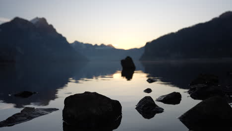 Time lapse of still lake at dusk with rocks in foreground and silhouette mountains in distance