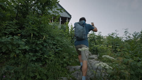 A-hiker-walking-on-a-path-uphill-towards-the-wooden-mountain-cottage-surrouden-with-green-plants