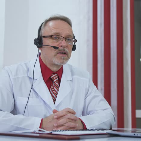 doctor with headset talking on camera in medical office. professional doctor in white medical coat sitting at desk and giving online consultation during quarantine.