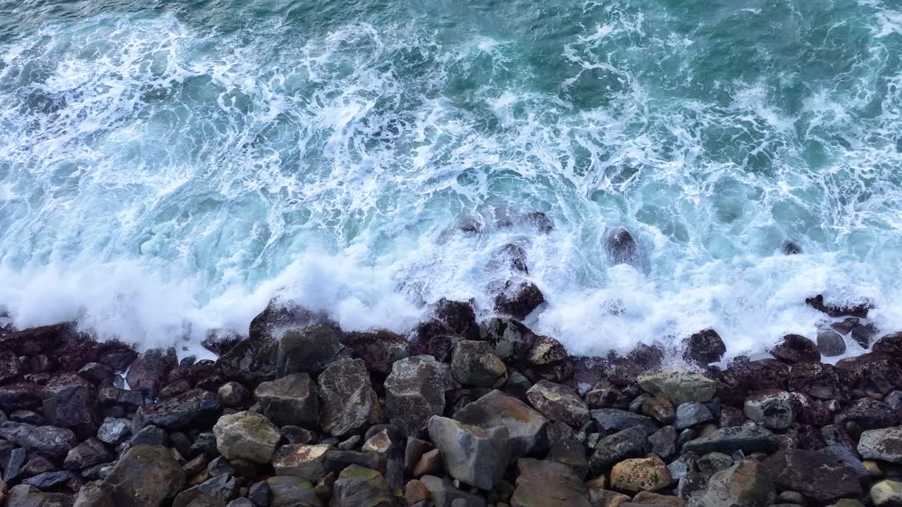 Aerial of waves smashing rocky Morro Beach shoreline near iconic Morro Rock in CA, textured ocean coastline backdrop