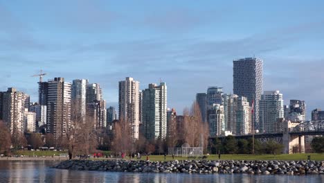 wide shot of a small park by the sea with people strolling by and highrise buildings in the background on a sunny winter day in vancouver, canada