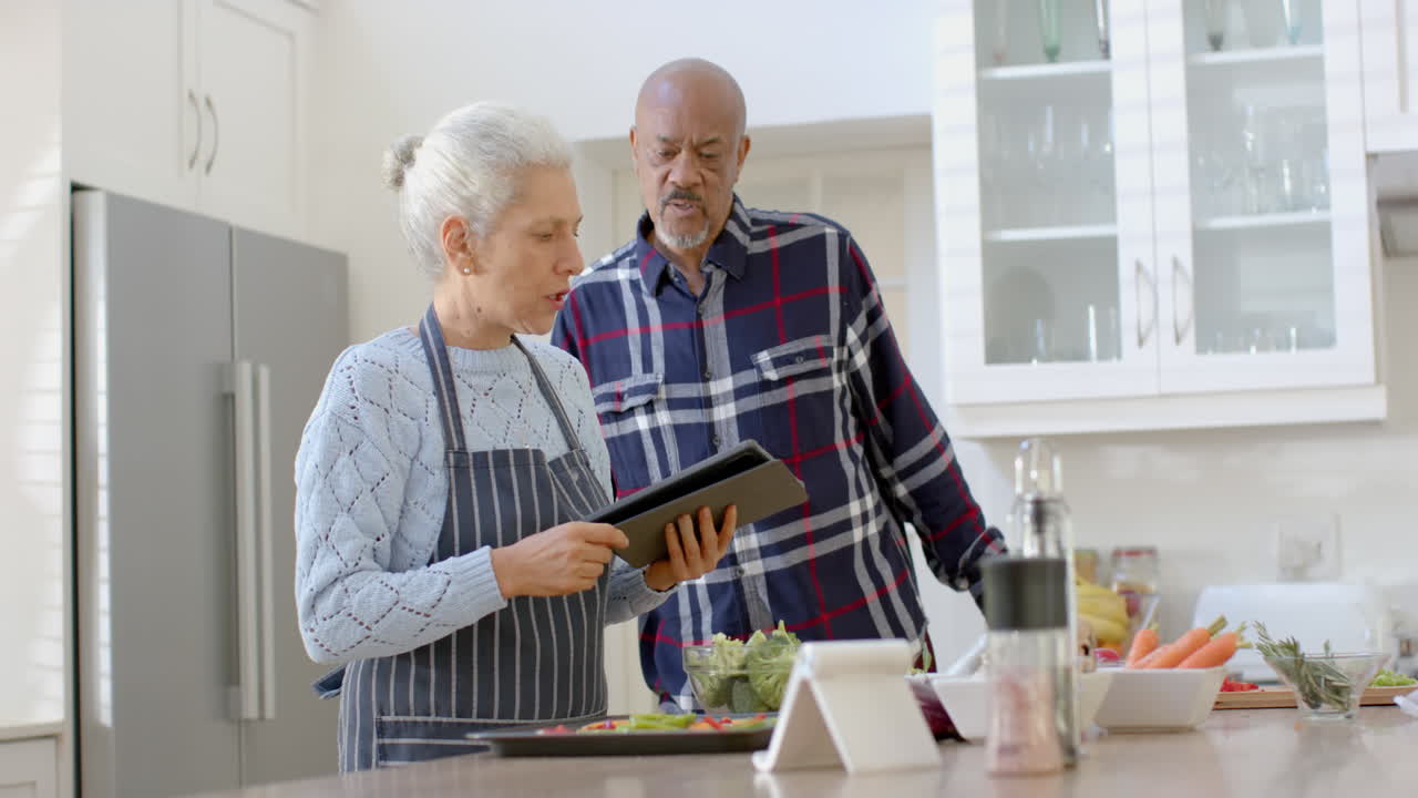una pareja biracial feliz usando una tableta y cocinando en la cocina, en cámara lenta
