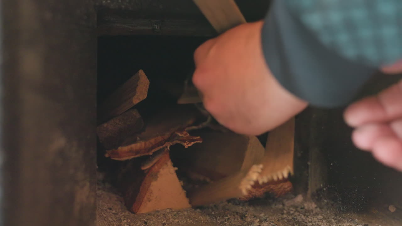Close up inside masonry chimney shows person using hand to wedge split fire wood pieces against sooty brick wall, preparing ignition, promising cozy warmth and ambience for winter evening