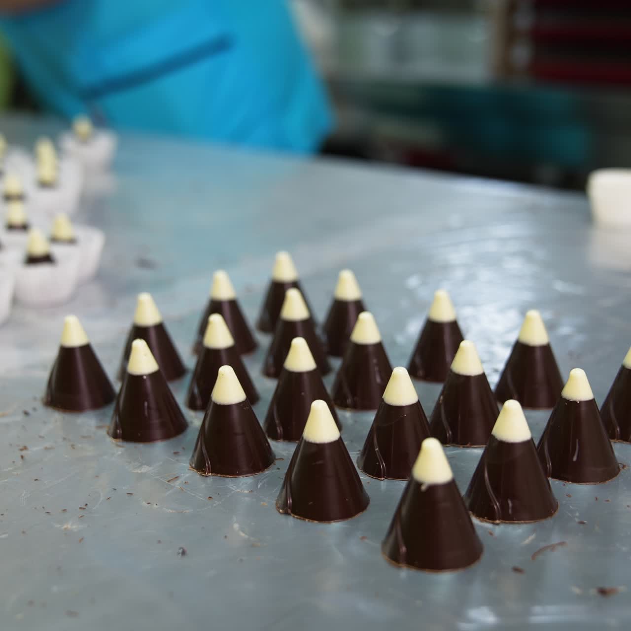 Confectionery worker turns the plastic mold the table. Cone chocolate candies with white tops are put on the metal surface. Candies packaging at backdrop