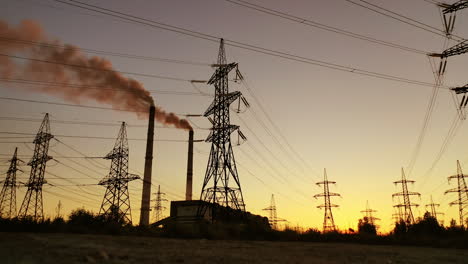 high voltage electric lines and industry. pipes with dark smoke near the transmission electric towers at dusk. pollution the atmosphere. view from below.