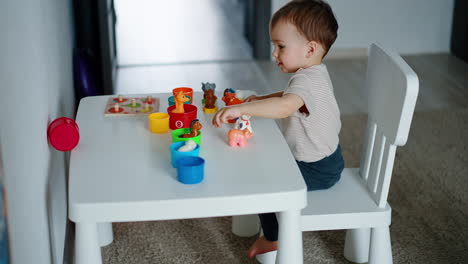 adorable baby plays with toys at the table. kid pushes the cup and watches it roll.