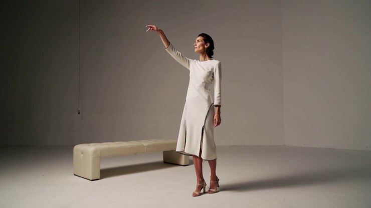 Partendo da una posa neutra, donna con vestito color crema che solleva il braccio destro verso l'alto in studio, con una panca a sinistra per il servizio fotografico