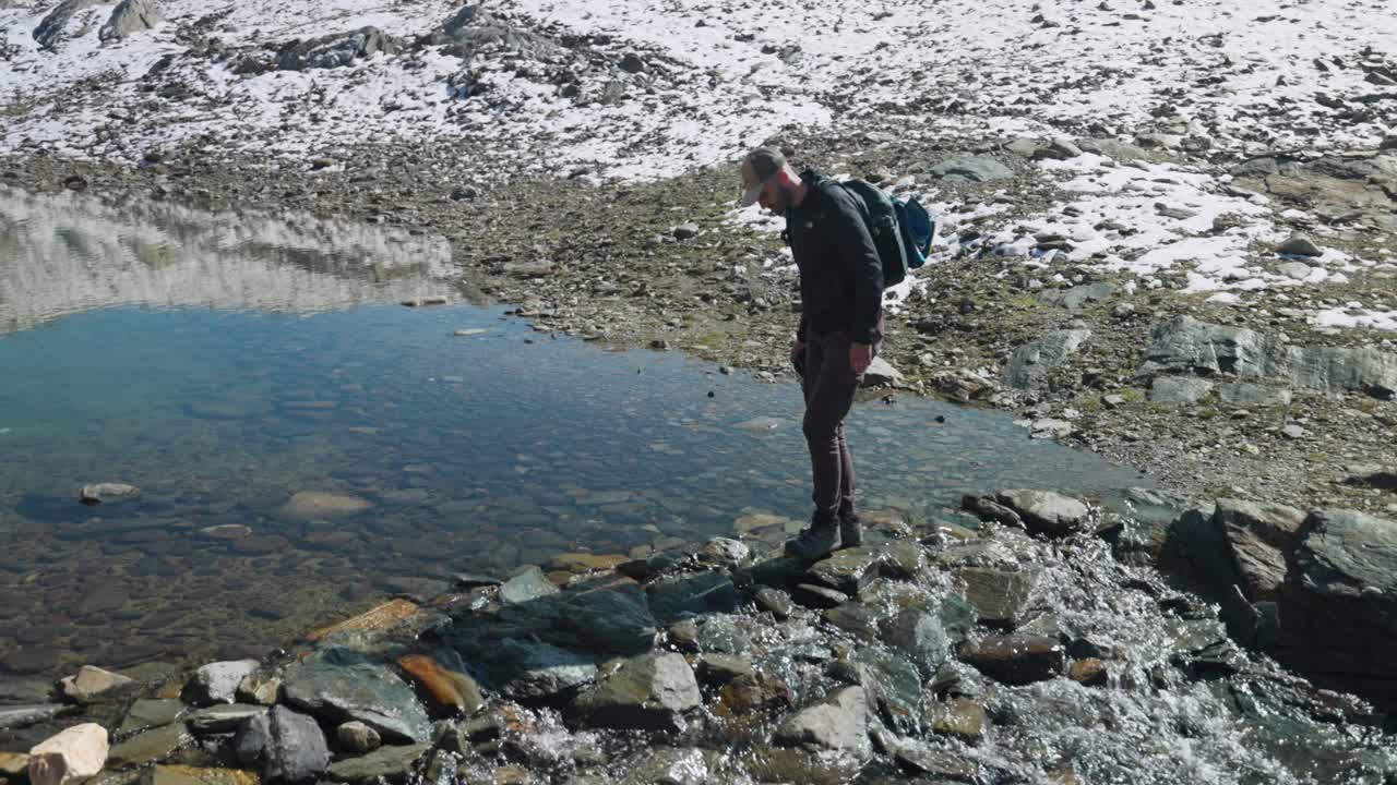 Solo Male Hiker Stepping Over Rocks Crossing Shallow Stream On Top Of Cima Fontana In Valmalenco